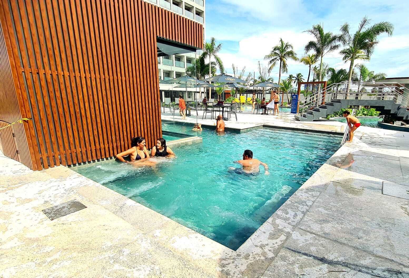 Une piscine où se baignent des personnes, entourée de palmiers et bordée d’un bâtiment à la façade en bois.
