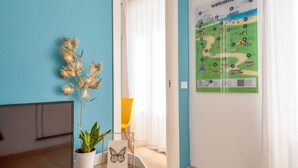 A room with a blue wall, a potted plant, a framed picture, a yellow chair, and a welcome poster. - The Echo in the Rock, Welcome Way (Vernazza)