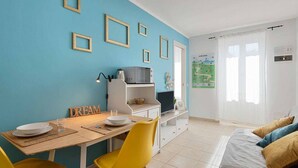 A room with a blue wall, a wooden dining table with yellow chairs, a bed with blue and yellow pillows, and a white door with a welcome sign. - The Echo in the Rock, Welcome Way (Vernazza)