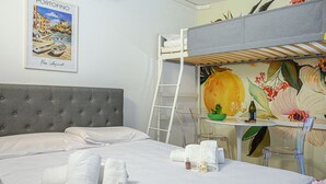 Una habitación de hotel con cama, literas, escritorio con una computadora portátil, un cuadro y artículos de aseo.