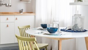 Ein Esstisch ist gedeckt mit blauen Schüsseln, einer klaren Glasgießkanne und einer weißen Gießkanne. Zwei Stühle, ein weißes Schrank und ein weißes Regal mit Dekorationen sind ebenfalls zu sehen.