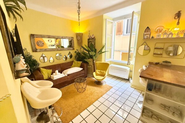 A cozy living room with a brown sofa, a yellow armchair, and a coffee table. There is a large mirror on the wall and a window with shutters.