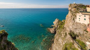 A coastal cliff with clear blue sea, a building with multiple windows, and a small structure on top. - Queen of Sea Diamond, Terre Marine (Manarola)