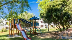 A playground with a slide and climbing structures, surrounded by trees and a building in the background.