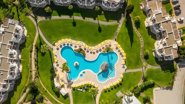 An aerial view of a resort with a central swimming pool, multiple lounge areas with umbrellas, and surrounding greenery.