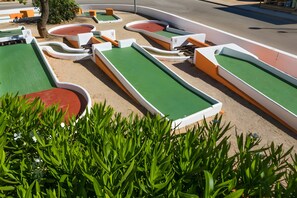 Um campo de mini golfe com vários buracos verdes e vermelhos, cercado por areia e limitado por estruturas brancas e laranjas.