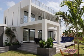 Une maison moderne à deux étages, dotée d’un balcon, de grandes fenêtres et d’un petit jardin agrémenté de plantes et d’un hamac.