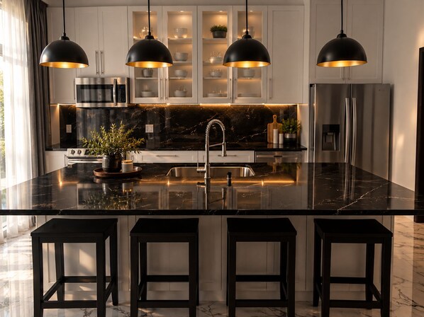 A modern kitchen with a central island, black pendant lights, and stainless steel appliances.