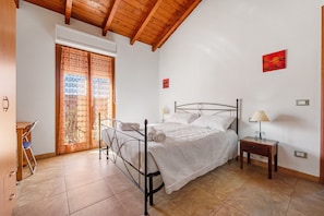 A bedroom with a bed, a desk, a chair, a nightstand with a lamp, and a window with curtains.