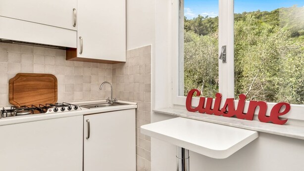 Une cuisine moderne avec des meubles blancs, une cuisinière à gaz et une fenêtre donnant sur un paysage verdoyant.