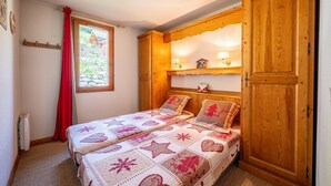 A bedroom with a bed, a window with red curtains, a wooden wardrobe, and a shelf with decorative items.