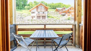 A wooden balcony with a table and two chairs, overlooking a stone wall and two wooden houses.