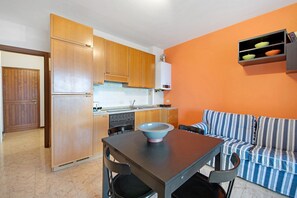 Una cocina compacta con armarios de madera, una mesa de comedor con sillas, un tazón azul sobre la mesa y un sofá a rayas frente a una pared naranja.