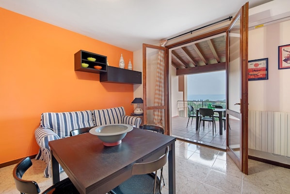 Une pièce comprenant une table à manger, un canapé, une petite étagère avec des objets décoratifs, un balcon avec vue et une porte donnant sur l’extérieur.