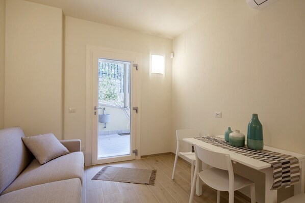 A compact living space with a sofa, a dining table with chairs, a small kitchen area, and a sliding glass door leading to an outdoor area.