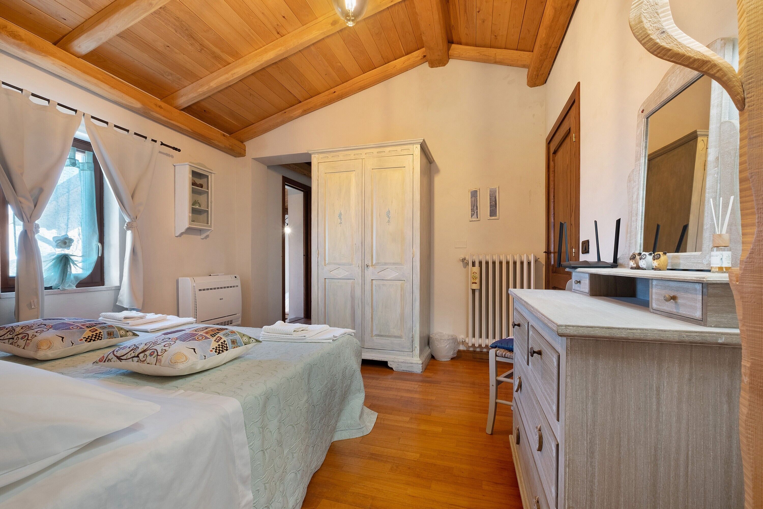 Un dormitorio con techo de madera, una cama con almohadones estampados, un armario, un radiador y un tocador con espejo.