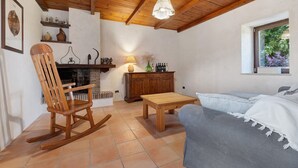 A cozy living room with a wooden ceiling, a fireplace, a rocking chair, a sofa, and a window with a view of greenery. - Room 'Among the Olive Trees and the View' with Mountain Views, Private Garden, and Wi-Fi (Orvieto)