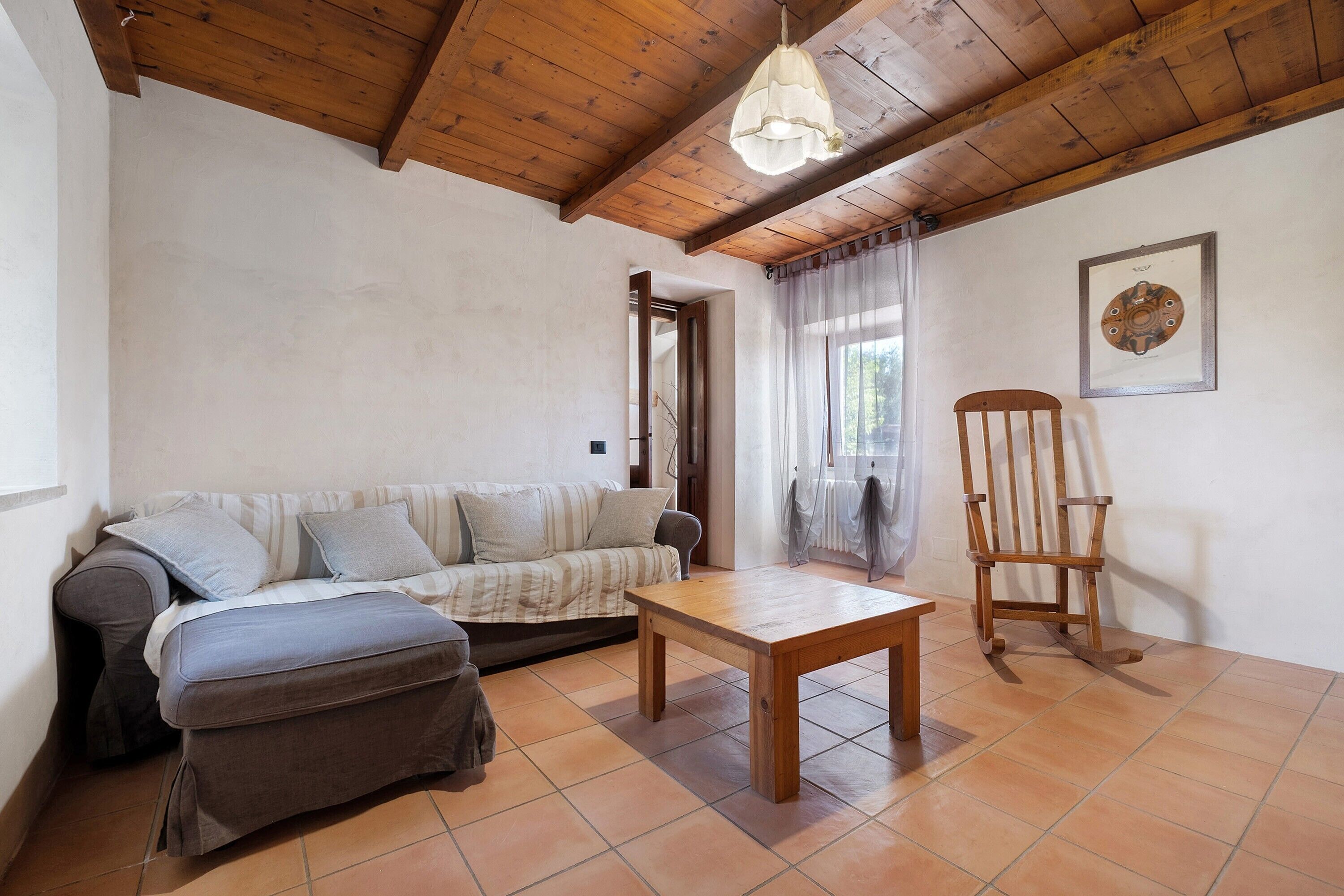 Una habitación con techo de madera, un sofá, una mesa, un sillón mecedora y un cuadro enmarcado en la pared.