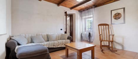 A room with a wooden ceiling, a sofa, a table, a rocking chair, and a framed picture on the wall.