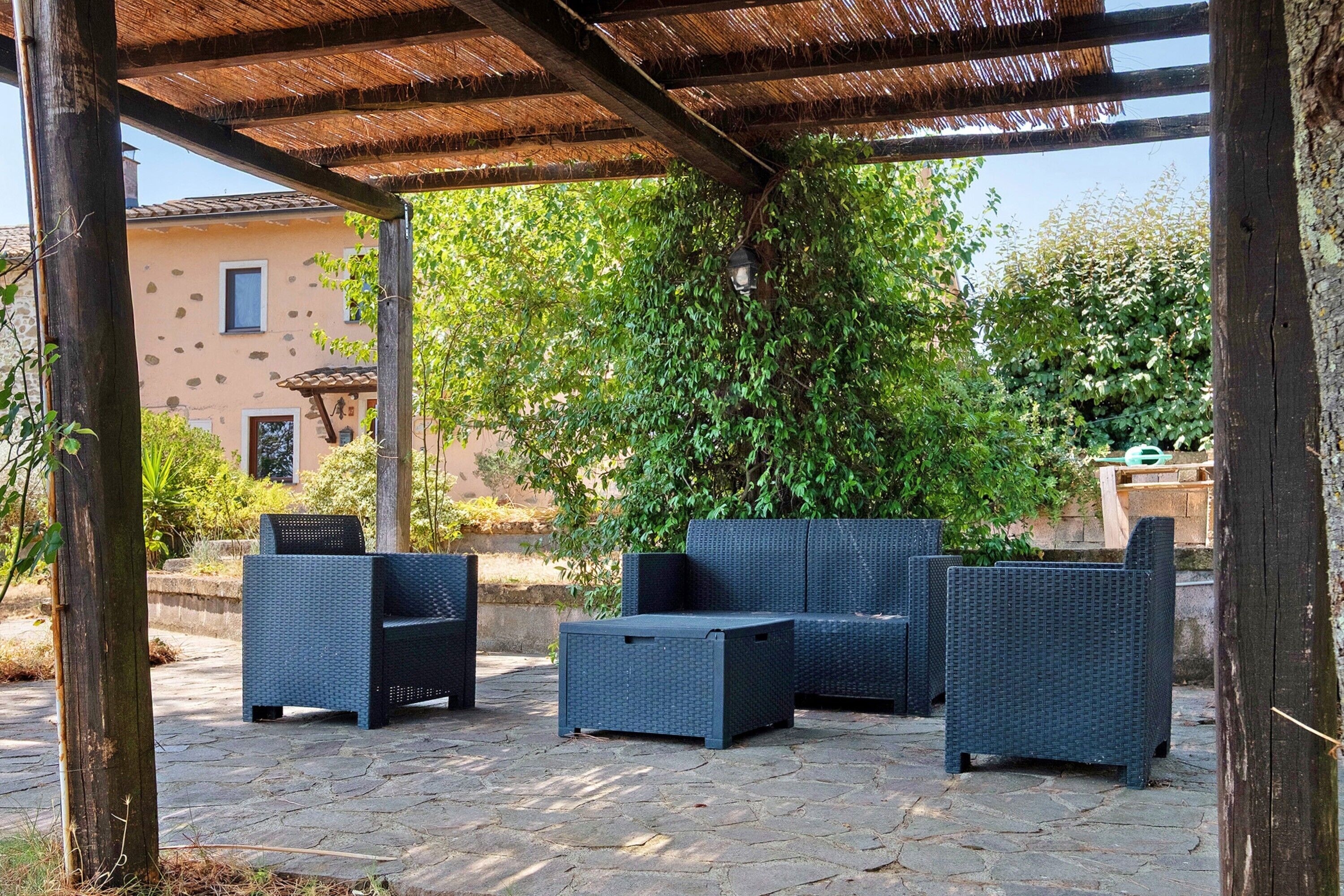 An outdoor seating area with wicker furniture under a pergola, surrounded by greenery and a building in the background.