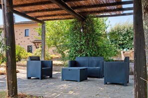Un área de estar al aire libre con mobiliario de mimbre bajo una pérgola, rodeada de vegetación y un edificio al fondo.