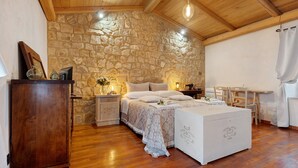A bedroom with a stone wall, wooden ceiling, a bed with white linens, a bedside table, a dresser, and a small desk with chairs. - Room 'Among the Olive Trees and the View' with Mountain Views, Private Garden, and Wi-Fi (Orvieto)