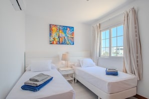 Quarto de hotel com duas camas, uma mesinha de cabeceira, um abajur, um quadro na parede e uma janela com cortinas.