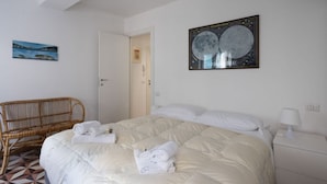 A neatly made bed with white bedding and rolled towels, a wicker chair, a framed picture of the moon, and a white nightstand with a lamp. - The Abode of the Poets, TerreMarine (Lerici)