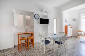 Une pièce comprenant une table à manger, des chaises, un téléviseur, un climatiseur et une fenêtre avec des rideaux.