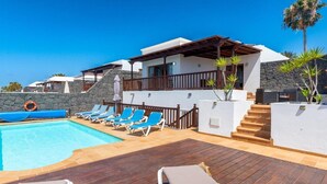 A poolside area with lounge chairs, a pool, and a building with a stone wall and wooden deck.