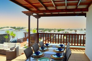 A covered outdoor dining area with a table set for four, surrounded by wicker chairs, overlooking a residential area with white buildings and greenery. - Vacation 'Casa Laura' with Heatable Pool, Sea View, Wi-Fi and Air Conditioning (Playa Blanca)