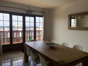 A dining area with a wooden table, white chairs, a bowl, large windows with a view, and a hanging light fixture. - Vacation 'Casa Laura' with Heatable Pool, Sea View, Wi-Fi and Air Conditioning (Playa Blanca)