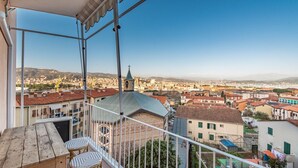A balcony with a wooden table and two wicker chairs, overlooking a cityscape with buildings and a church.