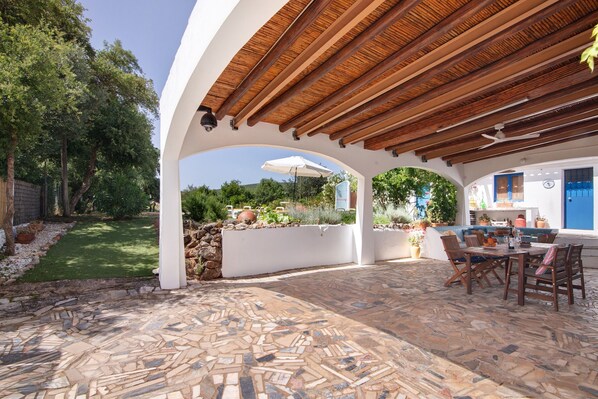 Portico esterno coperto con pavimento in pietra, arredi in legno e vista su giardino e casa.