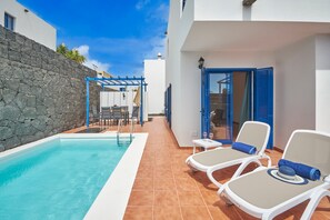 Área à beira da piscina com parede de pedra, pérgola, cadeiras de descanso e porta de vidro que dá para um quarto.