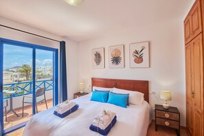 Quarto de hotel com uma cama, duas almofadas, mesa de cabeceira, luminária, janela com vista e obras de arte na parede.
