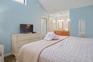 A bedroom with a bed, a television on a dresser, a bathroom with a mirror and sink, and a door leading to another room.