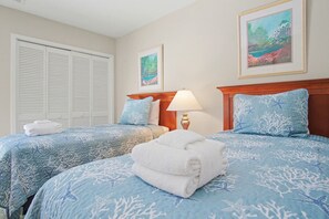 Quarto de hotel com duas camas, cada uma com colcha azul estampada com corais e estrelas-do-mar, toalhas brancas e cabeceira de madeira. Há duas pinturas emolduradas nas paredes, uma retratando uma paisagem e a outra uma cena de praia.