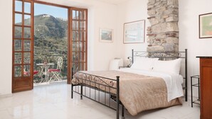 Un dormitorio con pared de piedra, una cama, balcón con mesa y sillas, y vista a una ladera.