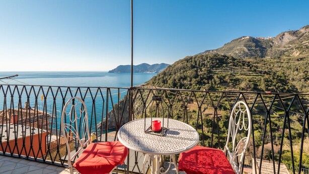Un balcón con una mesa redonda y cojines rojos, con vistas a un paisaje costero con colinas y un mar azul cristalino.
