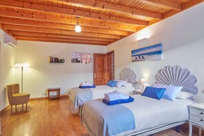 Una stanza con travi in legno al soffitto, due letti con lenzuola blu, una sedia, un tavolino e una mensola a muro con dei libri.