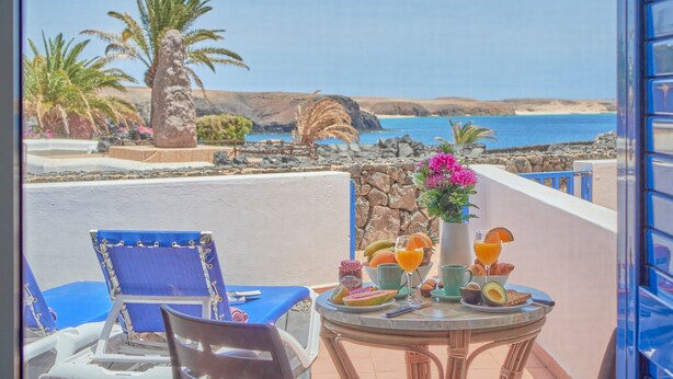 Een balkon met een ontbijttafel, uitkijkend over een kustlandschap met palmbomen en uitzicht op zee.