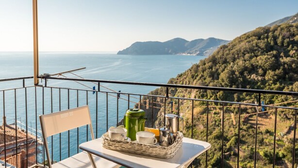 Un balcón con una mesa dispuesta para dos, con vista a un paisaje costero que incluye montañas y el mar.