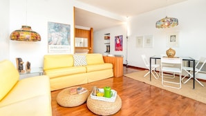 Una sala de estar luminosa con un sofá amarillo, mesas redondas y una zona de comedor con una mesa de cristal y sillas blancas.