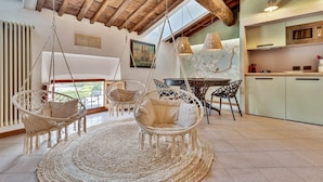 A cozy room with a high ceiling, rustic wooden beams, a round woven rug, two hanging wicker chairs, a dining table with chairs, a kitchen area with cabinets, and a radiator. - Magnolia, Welcome Way (Lerici)