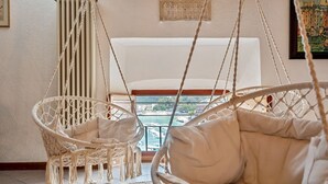 A room with two white wicker chairs with cushions, a woven rug, and a view of a body of water through a window. - Magnolia, Welcome Way (Lerici)