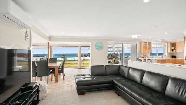 A modern living room with a black leather sofa, a dining area with a table and chairs, a kitchen with wooden cabinets, and a large window offering a view of the sea.