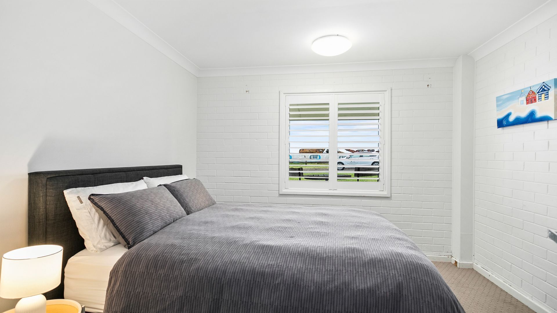 A bedroom with a bed, a window with blinds, a lamp, and a painting on the wall.