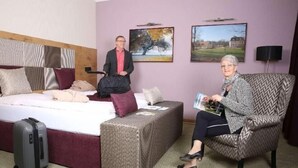 Hotellihuone, jossa on sänky, oleskelualue, matkalaukku ja kaksi ihmistä.