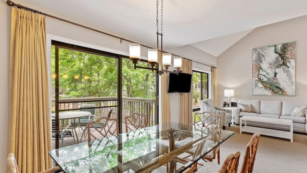 Une pièce spacieuse avec une grande table à manger en verre, des chaises en osier, un canapé blanc et un téléviseur fixé au mur.